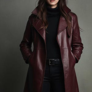 Könnte beinhalten: Ein burgunderfarbener Ledertrenchcoat mit Reverskragen und passendem Gürtel. Der Mantel wird über einem schwarzen Rollkragenpullover und einer schwarzen Hose getragen. Das Model steht vor einem grauen Hintergrund.