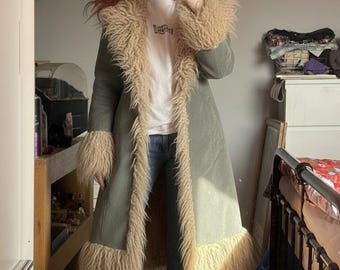 May include: A long, sage green coat with tan faux fur trim on the collar, cuffs, and front edges. The coat is open, revealing a white t-shirt and blue jeans. The coat has a vintage, bohemian style.