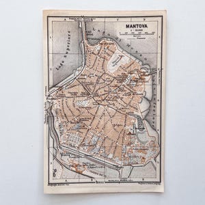 Op de afbeelding: Een vintage zwart-wit gedrukte kaart van Mantua, Italië, met beige gekleurde stedelijke gebieden, gedetailleerde stratenplannen en de omliggende meren Lago Superiore en Lago Inferiore. De kaart bevat een schaal, rasterlijnen en tekst.