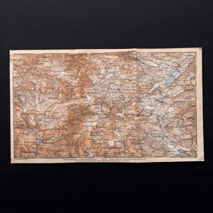 Op de afbeelding: Een antieke kaart met een gedetailleerd topografisch ontwerp, met bruine en blauwe tinten. De kaart toont geografische kenmerken, waaronder bergen, rivieren en meren. De kaart is gedrukt op verouderd papier, met zichtbare slijtage.