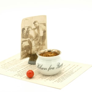 May include: A white ceramic pot with a gold rim and the text "Hilsen fra Berg" in black script. The pot is sitting on a white surface with a red marble and a vintage photograph in the background.