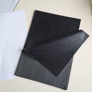 May include: A stack of rectangular black tissue paper sheets with a visible crinkled texture. One sheet is folded over at an angle, revealing the matte surface and the thin, semi-translucent quality of the paper. A single white sheet is visible.