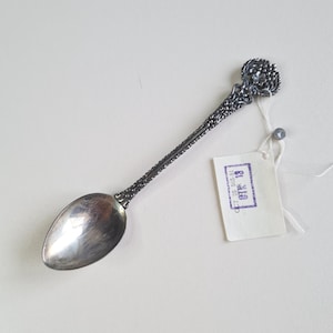 Op de afbeelding: Een zilverkleurige metalen lepel met een gedecoreerd, gestructureerd handvat met een druiventrosmotief aan de bovenzijde. Een wit papieren label is met een touwtje aan het handvat bevestigd, met de paarse stempeltekst OCT 25 565-81 QTK 18.