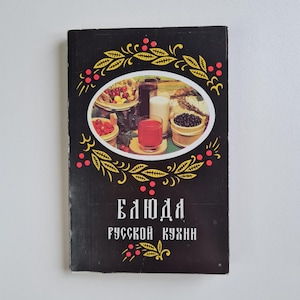 Könnte beinhalten: Ein schwarzes Kochbuch mit dem Titel "БЛЮДА РУССКОЙ КУХНИ" und einer zentralen ovalen Abbildung von Beeren, Holzschalen und Gläsern mit Saft. Der Einband ist mit einem gelben und roten Blatt- und Beerenmuster um das Bild herum verziert.