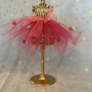 May include: A decorative gold-colored dress form with a red tulle skirt and bow. Small rose accents are attached to the skirt. The dress form is on a gold-colored base and stand.