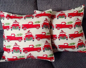 May include: Two beige throw pillows with a red truck pattern. The trucks are carrying Christmas trees and have a small house on top. The background is a beige color with white dots.