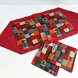 May include: A red quilted table runner with a patchwork design of red, green, blue, and yellow squares. The runner is accented with hearts, stars, and plaid patterns. Two matching quilted coasters are included.