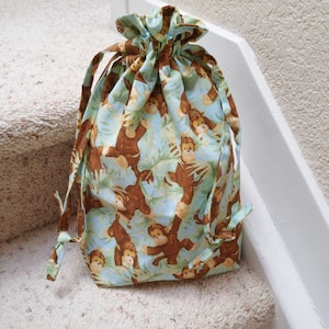 May include: A drawstring bag made of light blue fabric with a pattern of brown monkeys on green leaves. The bag has a gathered top and long ties.