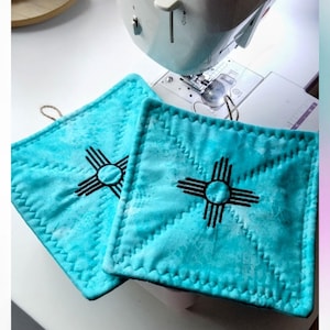 May include: Two square, turquoise fabric pot holders featuring a black embroidered Zia sun symbol in the center. Each piece has decorative zigzag stitching along the edges and a small brown twine loop attached to the top corner for hanging.