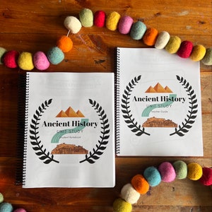 May include: Two spiral bound notebooks with white covers and black text. The covers feature a black and white illustration of a wreath with the text "Ancient History Unit Study" and the words "Student Notebook" and "Teacher Guide". The notebooks are on a wooden surface with a colorful garland of felt balls.
