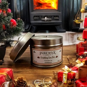 Op de afbeelding: Een zilveren kaars pot met een zwart-wit label waarop staat "purewick candle co. crackling log fire 23 hour burn time homemade vegan soy wax". De kaars staat op een houten oppervlak met een open haard op de achtergrond.