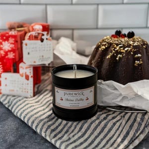 Könnte beinhalten: Ein schwarzes Kerzengefäß mit einem weißen Etikett, auf dem "Purewick Candles Co. Christmas Pudding 27 Hour Burn Time Homemade" steht. Die Kerze steht auf einem weiß-blau gestreiften Stoff.