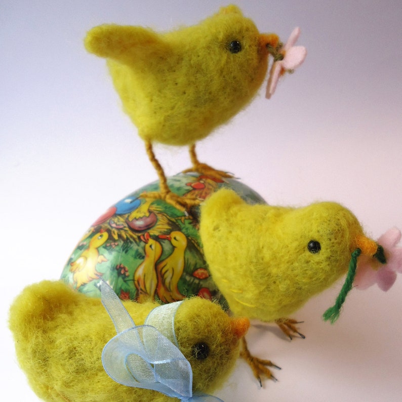 May include: Three yellow felt chicks, each with black eyes and orange beaks, are arranged around a decorated egg. One chick holds a pink flower. The egg is painted with a pastoral scene of ducks and other birds.