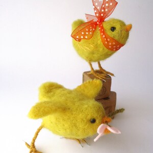May include: Two handmade yellow felt chicks. One chick is standing on a wooden block and has an orange polka dot bow. The other chick is lying down and holding a pink flower. The chicks have black eyes and orange beaks.