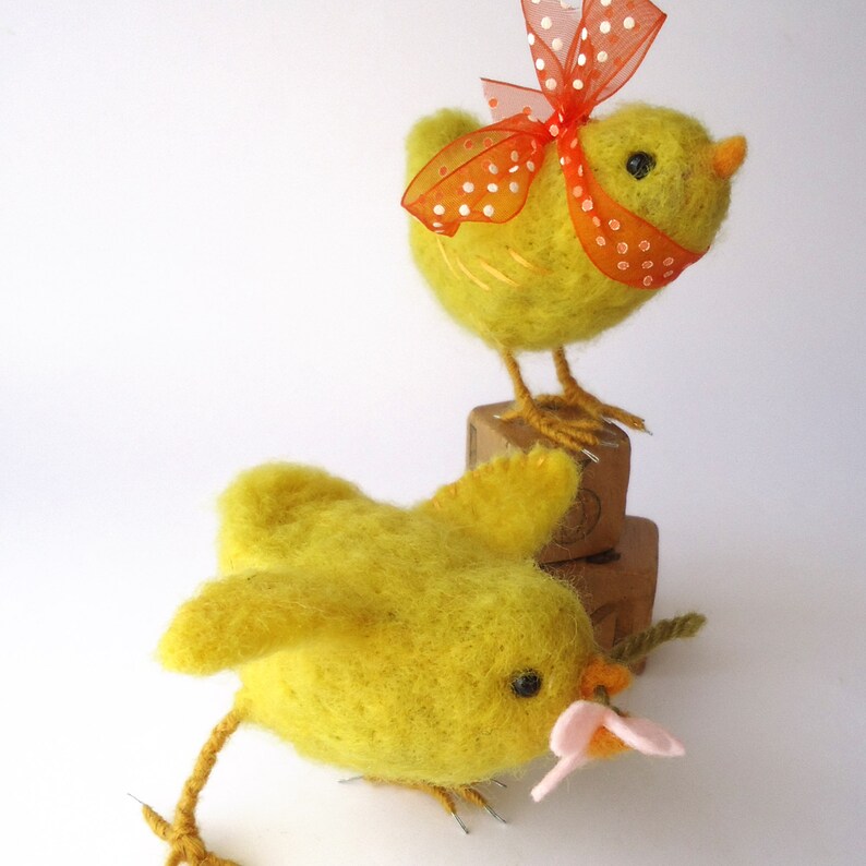 May include: Two handmade yellow felt chicks. One chick is standing on a wooden block and has an orange polka dot bow. The other chick is lying down and holding a pink flower. The chicks have black eyes and orange beaks.