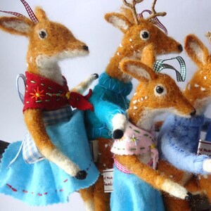 K&ouml;nnte beinhalten: Vier handgefertigte Filz-Hirsche, jede mit einem anderen Outfit. Ein Hirsch tr&auml;gt einen blauen Pullover, ein anderer einen rosa-wei&szlig;en Schal und ein anderer einen rot-wei&szlig;en Schal. Der vierte Hirsch tr&auml;gt ein blaues Kleid mit einem wei&szlig;en und blauen Gingham-Muster.