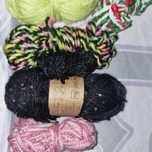 Könnte beinhalten: Eine Sammlung bunter Wollknäuel in verschiedenen Farbtönen, darunter Limettengrün, Schwarz mit Pailletten, Rosa und Lavendel. Das Bild zeigt verschiedene Texturen und Muster, ideal für Strick- oder Häkelprojekte.