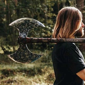 Könnte beinhalten: Eine Person hält eine große doppelseitige Axt mit einem Holzstiel und Lederumwicklungen. Die Axt hat eine silberne Klinge mit einer gehämmerten Oberfläche und der Stiel ist mit Runen beschriftet.