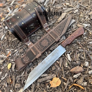 Puede incluir: Un cuchillo de hoja plateada grande con un mango de madera y runas grabadas, exhibido con una funda de cuero marrón y un contenedor de cuero a juego. La funda presenta un diseño de lobo y patrones intrincados. El logotipo de War Blades es visible.