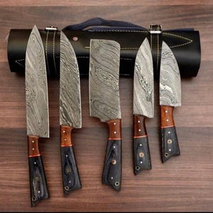 May include: Set of five kitchen knives with patterned blades and wooden handles. The knives are arranged in a row on a wooden surface and are rolled up in a black leather case.