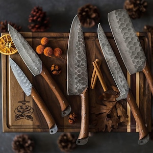 May include: A collection of six kitchen knives with wooden handles and patterned steel blades, arranged on a wooden cutting board. The knives vary in size and shape, with a mix of textures and designs. The scene is decorated with cinnamon sticks, dried orange slices, and pine cones.