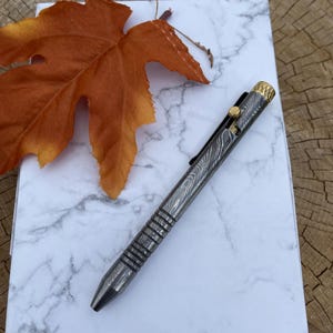 May include: A Damascus steel pen with a gold-colored clip and accents rests on a marble-patterned notepad. An orange leaf is placed near the pen. The pen has a textured, patterned design.