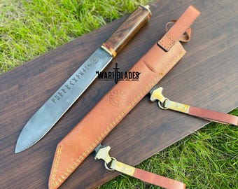 May include: A long, silver blade knife with a wooden handle and etched runes, resting on a wooden surface. A brown leather sheath and two leather straps with metal clasps are also present. The knife is labeled "WARH BLADES".
