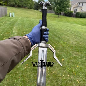 May include: A silver sword with a black wrapped handle and a detailed wolf head pommel. The blade has a central fuller and the text "WARH BLADES" is visible. The sword is held against a green grassy background.