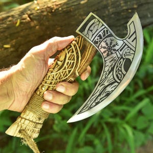 May include: A hand-held, silver-bladed axe with a wooden handle. The handle is intricately carved with geometric patterns and wrapped with twine. The axe head features etched designs, including a snowflake-like symbol. The axe is held against a blurred green background.