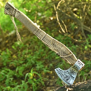 May include: A handcrafted axe with a wooden handle and a metal blade. The handle is wrapped with twine and features intricate carvings. The blade has a detailed design with a silver finish. The axe is a rustic, decorative item.