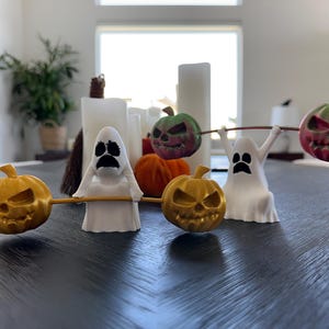 Op de afbeelding: Halloween-thema decoraties met geesten en pompoenen. Twee witte spookfiguren tillen gouden en groene pompoengewichten op. De pompoenen hebben gesneden gezichten. Andere pompoenen en kaarsen staan op de achtergrond.