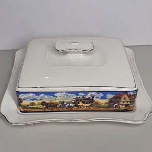 May include: A rectangular white ceramic butter dish with a matching lid featuring a gold-trimmed handle. The base of the lid is decorated with a colorful scene of a horse-drawn carriage traveling past a building under a blue sky with clouds.