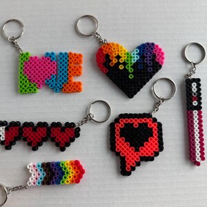 May include: A collection of colorful keychains made from plastic beads. Designs include the word "LOVE", a rainbow heart, a series of hearts, a rainbow arrow, a speech bubble with a heart, and a vertical design. Each keychain has a silver ring.