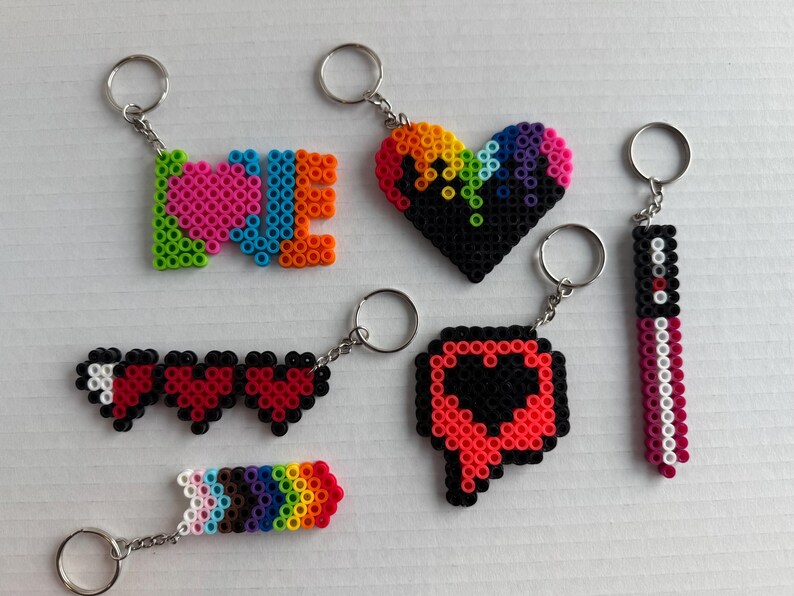 May include: A collection of colorful keychains made from plastic beads. Designs include the word "LOVE", a rainbow heart, a series of hearts, a rainbow arrow, a speech bubble with a heart, and a vertical design. Each keychain has a silver ring.