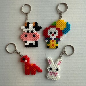 May include: Four handmade keychains featuring animal designs. Includes a black and white cow, a clown with a colorful balloon, a red dinosaur, and a white bunny with pink accents. Each keychain has a silver ring.