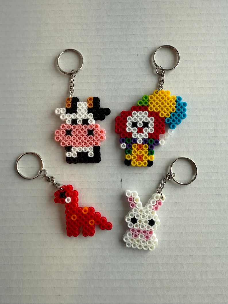 May include: Four handmade keychains featuring animal designs. Includes a black and white cow, a clown with a colorful balloon, a red dinosaur, and a white bunny with pink accents. Each keychain has a silver ring.