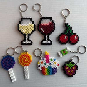 May include: A collection of handmade keychains featuring various designs. Includes pixelated wine glasses, cherries, lollipops, a cupcake, a piece of candy, and a strawberry. Each keychain has a silver metal ring for attaching to keys or bags.