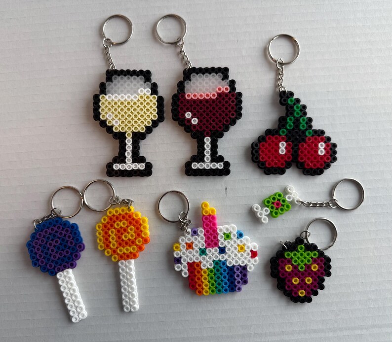May include: A collection of handmade keychains featuring various designs. Includes pixelated wine glasses, cherries, lollipops, a cupcake, a piece of candy, and a strawberry. Each keychain has a silver metal ring for attaching to keys or bags.