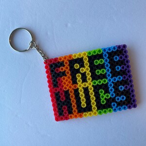 May include: A rectangular keychain made of colorful plastic beads. The keychain features the word "PRIDE" in yellow letters against a black background, framed by a rainbow border. A silver metal keyring is attached.