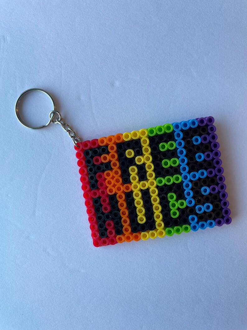May include: A rectangular keychain made of colorful plastic beads. The keychain features the word "PRIDE" in yellow letters against a black background, framed by a rainbow border. A silver metal keyring is attached.