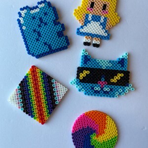 May include: A collection of colorful pixel art creations. Includes a blue square, a character resembling Alice in Wonderland, a blue cat with a mask, a rainbow diamond, and a rainbow spiral. Each piece is made of small, colorful beads.