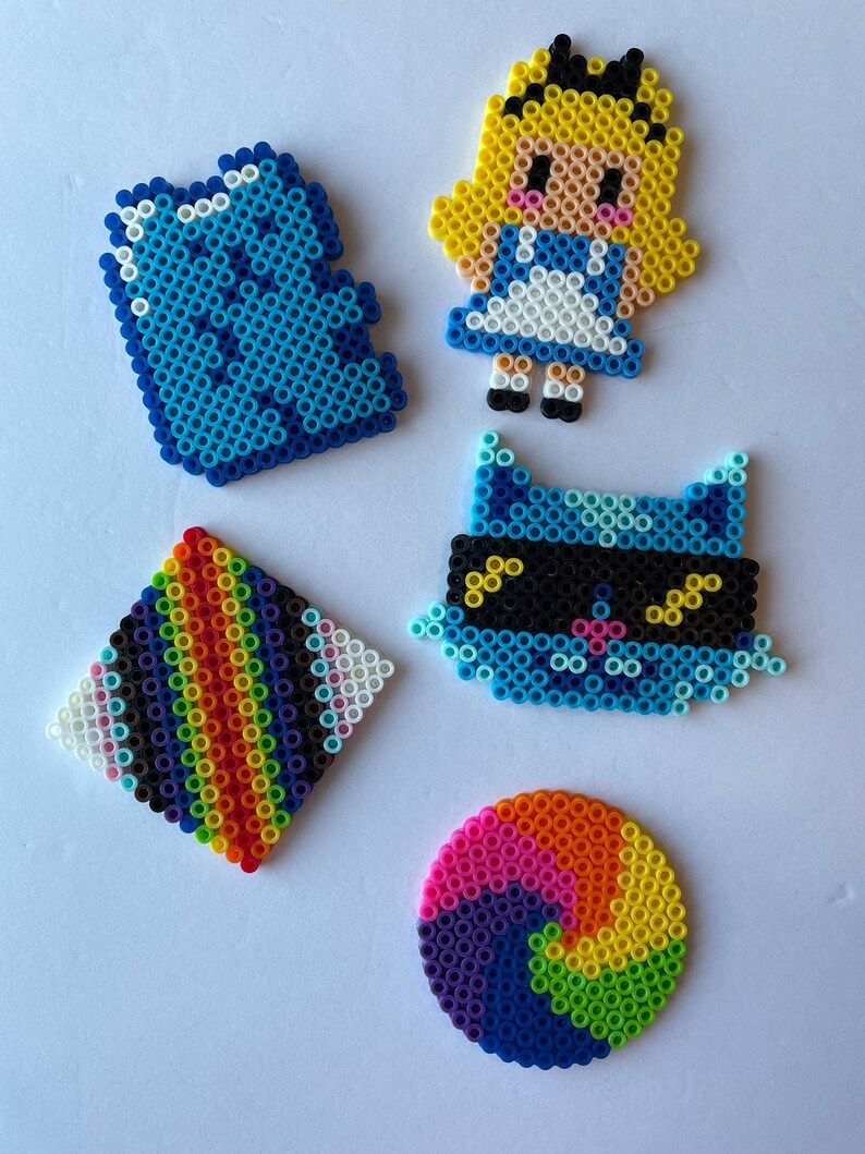 May include: A collection of colorful pixel art creations. Includes a blue square, a character resembling Alice in Wonderland, a blue cat with a mask, a rainbow diamond, and a rainbow spiral. Each piece is made of small, colorful beads.