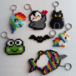 May include: A collection of handmade keychains featuring various colorful designs. Includes a rainbow-colored pony, a penguin holding a heart, a black cat, a green frog, a bat, a unicorn, and a rainbow fish, all made with small beads.