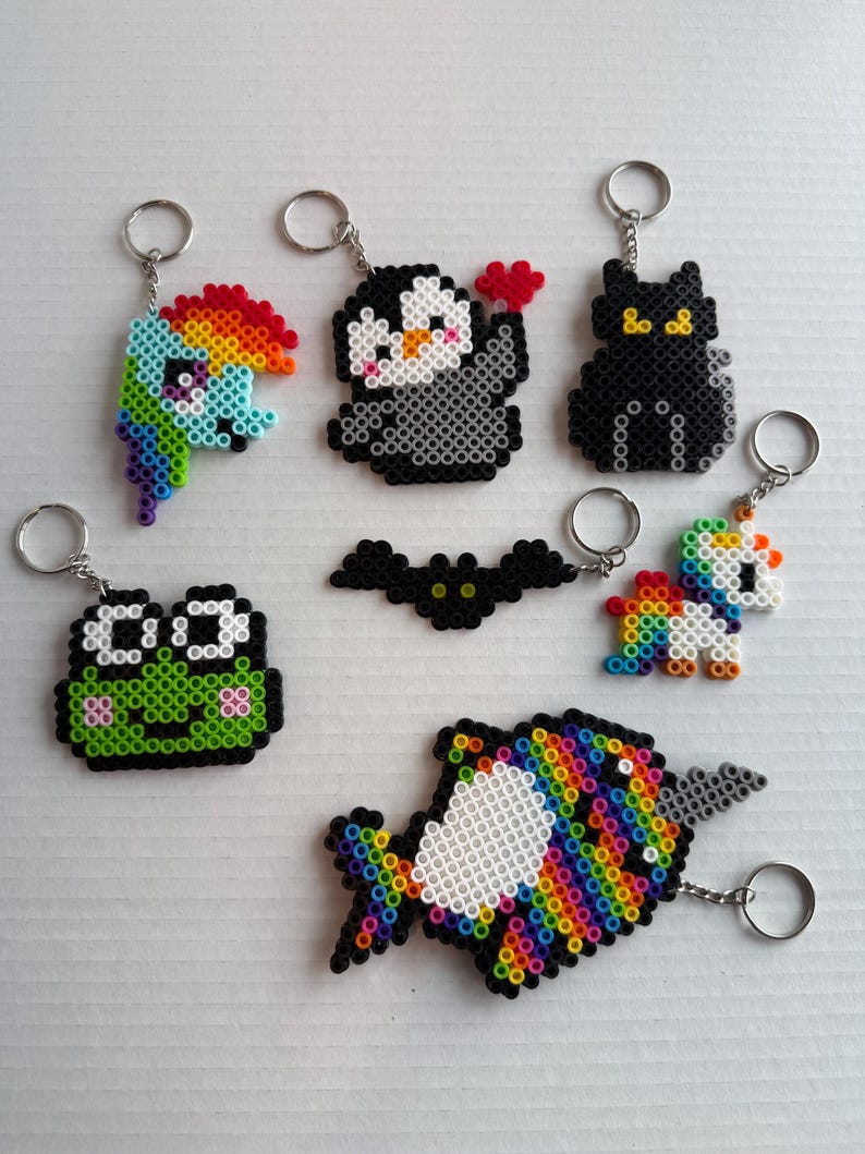 May include: A collection of handmade keychains featuring various colorful designs. Includes a rainbow-colored pony, a penguin holding a heart, a black cat, a green frog, a bat, a unicorn, and a rainbow fish, all made with small beads.
