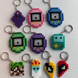 May include: A collection of colorful pixel art keychains. Designs include a purple diamond, pink and blue pixelated devices, a green square, a purple device, a teal square, a Minecraft-inspired block, a crowned figure, and two halves of a heart.