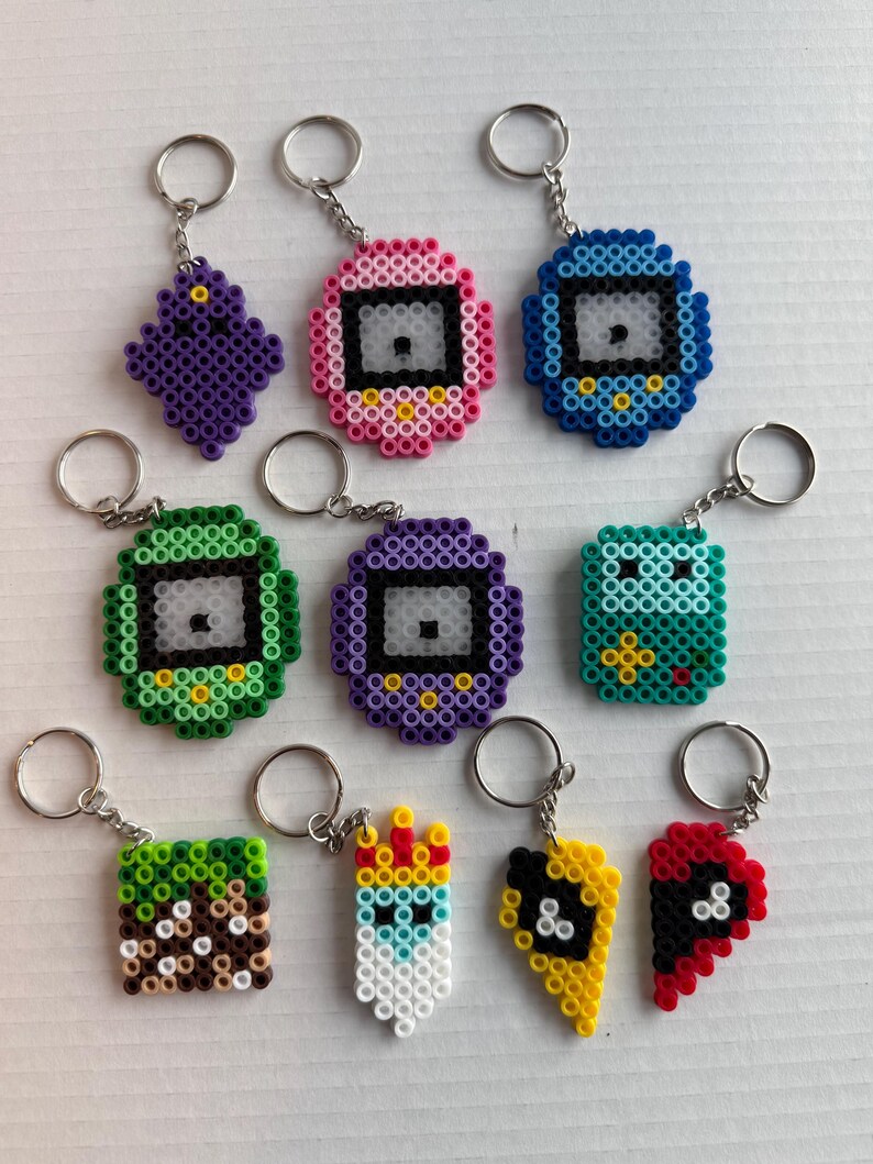 May include: A collection of colorful pixel art keychains. Designs include a purple diamond, pink and blue pixelated devices, a green square, a purple device, a teal square, a Minecraft-inspired block, a crowned figure, and two halves of a heart.
