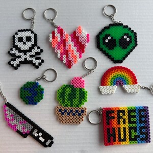 May include: A collection of handmade keychains in various shapes and colors. Designs include a skull, heart, alien, rainbow, knife, globe, cactus, and a rectangle with the text "FREE HUGS". Each keychain has a silver-colored ring.