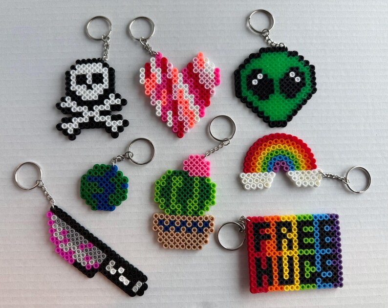 May include: A collection of handmade keychains in various shapes and colors. Designs include a skull, heart, alien, rainbow, knife, globe, cactus, and a rectangle with the text "FREE HUGS". Each keychain has a silver-colored ring.