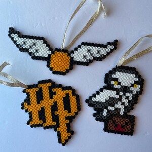 May include: Three handmade ornaments in a Harry Potter theme. One is a golden snitch with white and black wings. Another is a black and gold HP logo. The third is a black and white owl with a brown package.