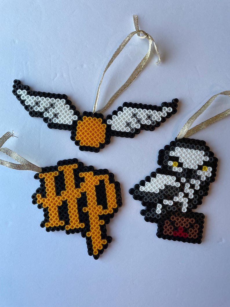 May include: Three handmade ornaments in a Harry Potter theme. One is a golden snitch with white and black wings. Another is a black and gold HP logo. The third is a black and white owl with a brown package.